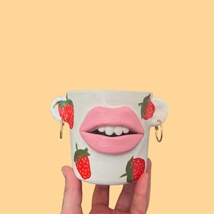 May include: White ceramic planter shaped like a face with a pink painted mouth and white teeth. The planter is decorated with red and green painted strawberries.
