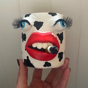 May include: A white ceramic planter with black cow spots, blue eyes, long eyelashes, and red lips holding a lit cigarette. The planter is a unique decorative item.