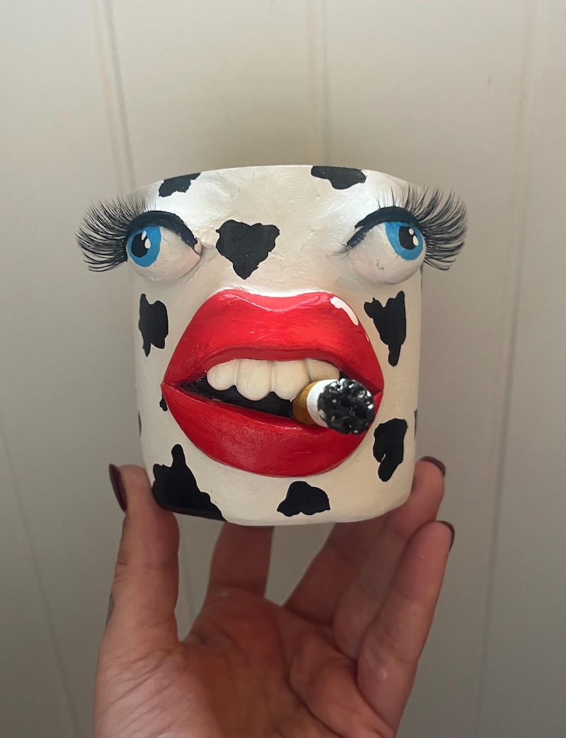 May include: A white ceramic planter with black cow spots, blue eyes, long eyelashes, and red lips holding a lit cigarette. The planter is a unique decorative item.