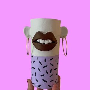 Pode incluir: Um vaso de cerâmica branco com um lábio castanho e dentes brancos, pintado com um padrão geométrico roxo e preto. O vaso tem brincos de argola dourados de cada lado.