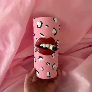 Puede incluir: Jarrón de cerámica rosa con un diseño de estampado de leopardo blanco y negro. El jarrón presenta una boca roja esculpida en 3D con dientes blancos y un cigarrillo en su interior.