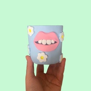 Puede incluir: Un macetero de cerámica azul claro con una boca rosa y dientes blancos. El macetero está decorado con margaritas blancas con centros amarillos.
