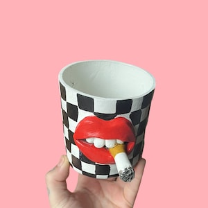 Puede incluir: Maceta de cerámica blanca, negra y roja con un patrón de tablero de ajedrez. La maceta presenta un par de labios rojos con un cigarrillo encendido. El interior de la maceta es blanco.