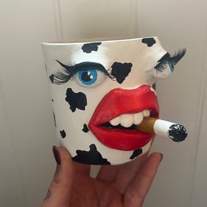 May include: A unique, hand-painted ceramic planter with a cow-like pattern. The planter features a face with blue eyes, long eyelashes, red lips, and a cigarette in its mouth. A creative and quirky decorative item.