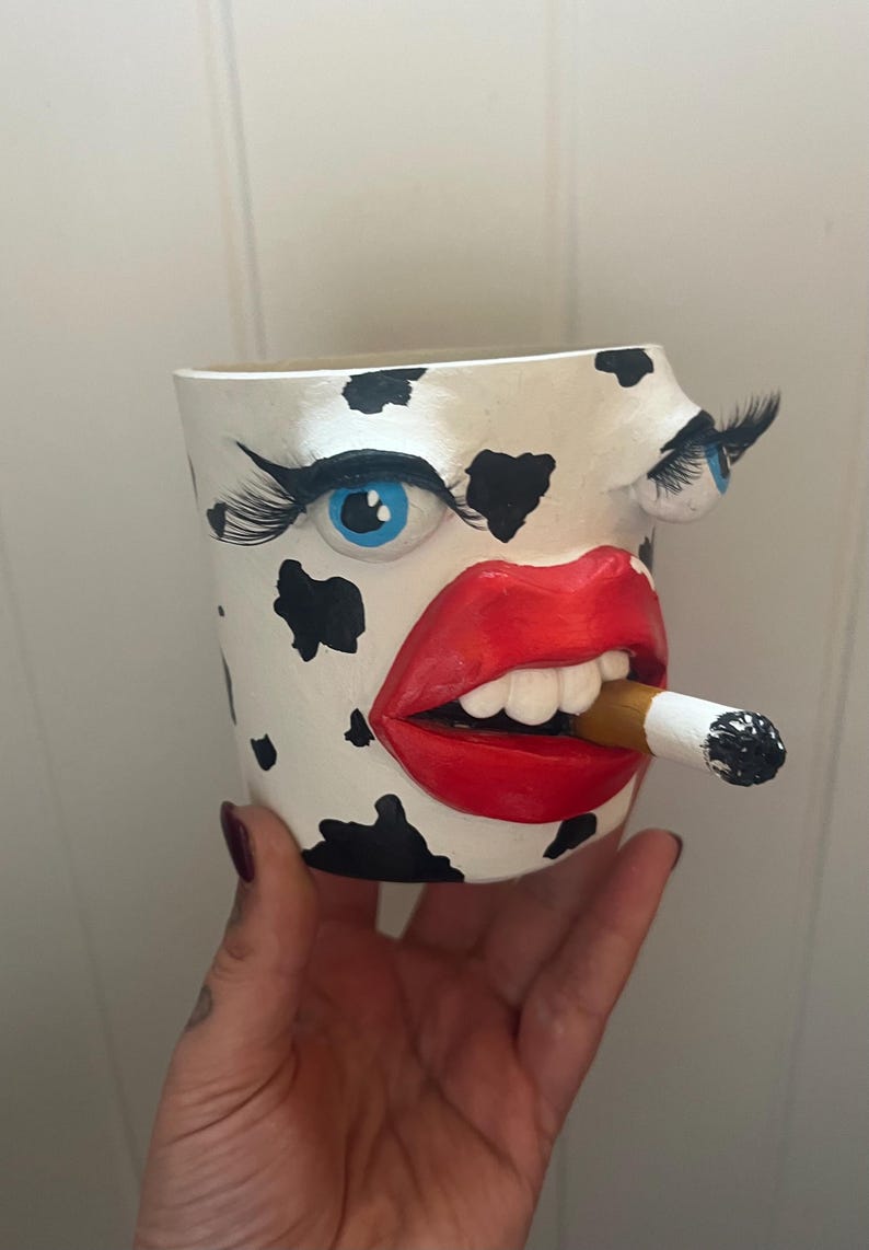 May include: A unique, hand-painted ceramic planter with a cow-like pattern. The planter features a face with blue eyes, long eyelashes, red lips, and a cigarette in its mouth. A creative and quirky decorative item.