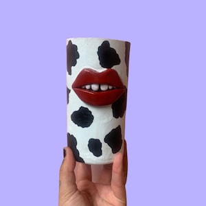 May include: A white ceramic cylinder vase with a black and white cow print design. A red sculpted mouth with white teeth is attached to the front of the vase.