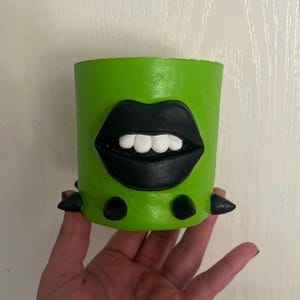 Puede incluir: Una maceta cilíndrica verde con un diseño de labios negros y dientes blancos. La maceta tiene púas negras alrededor de la base. La maceta está sostenida por una mano.