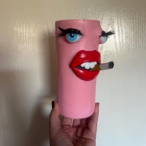 May include: A pink ceramic vase with a painted face, blue eyes, red lips, and a cigarette in its mouth.