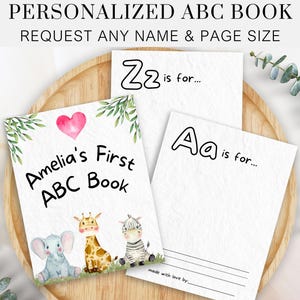 Może przedstawiać: Spersonalizowana książka ABC z akwarelową ilustracją żyrafy, zebry i słonia. Okładka zawiera tekst "Amelia's First ABC Book" z różowym sercem. Książka jest wykonana z miłością i można ją spersonalizować dowolnym imieniem i rozmiarem strony.