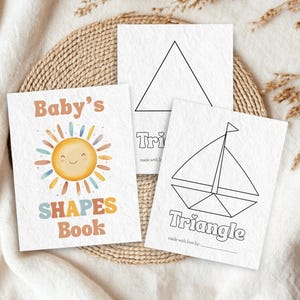 May include: A children's shapes book with a smiling sun illustration and the text "Baby's Shapes Book." Two additional cards show a triangle and a sailboat, both labeled "Triangle." The cards are on a woven mat.