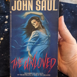 May include: A paperback book cover with a blue background featuring a woman with a skull face wearing a blue dress. The title of the book is "The Unloved" in red letters. The author is John Saul. The text below the title reads "The horrifying new novel by the bestselling author of The Unwanted."