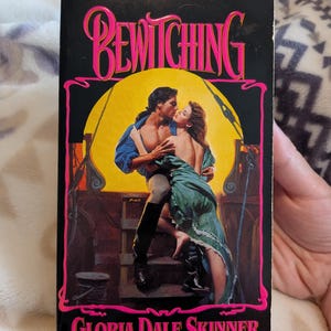 May include: A vintage paperback book titled "Bewitching" by Gloria Dale Skinner. The cover features a romantic illustration of a couple embracing, set against a yellow backdrop. The title and author's name are in pink lettering.