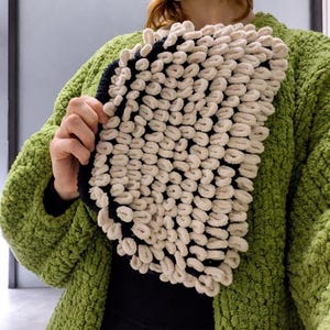 May include: A cream-colored, textured scarf with a black border is held up against a green textured cardigan. The scarf has a looped design, creating a soft, plush appearance. The cardigan is a vibrant green color.