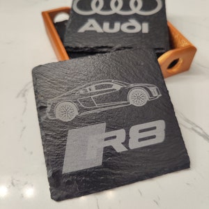 May include: Black slate coasters with a silver Audi logo and the text "Audi" on one coaster and a silver Audi R8 car and the text "R8" on the other coaster.