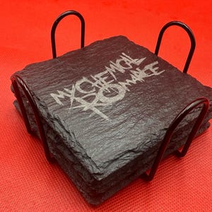 Puede incluir: Un juego de cuatro posavasos cuadrados de pizarra con el nombre de la banda "MY CHEMICAL ROMANCE" grabado en blanco. Los posavasos están apilados en un soporte de metal negro, sobre un fondo rojo.