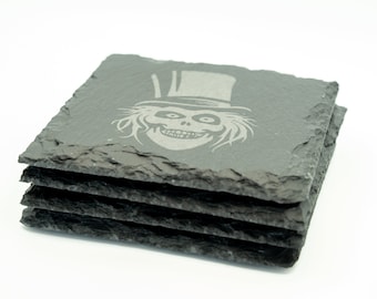 May include: Set of four black slate coasters featuring a gray engraved image of a ghost wearing a top hat.