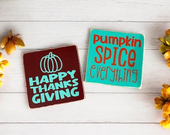 May include: Two wooden signs with fall-themed text. One sign is maroon with a pumpkin graphic and the words "HAPPY THANKS GIVING" in teal. The other is teal with the words "pumpkin SPICE everything" in orange.