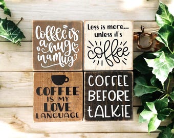 May include: Four wooden signs with coffee-themed sayings. The signs read "Coffee is a huge in a mug", "Less is more... unless it's coffee", "Coffee is my love language", and "Coffee before talkie".