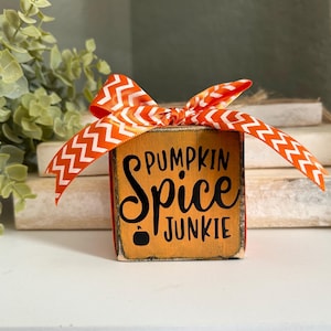 May include: A wooden block with the words "PUMPKIN SPICE JUNKIE" in black script, topped with an orange and white chevron ribbon. The block has an orange background and a small black pumpkin graphic. The item is a fall-themed decoration.