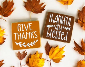 May include: Two square wooden signs with white lettering and fall leaves. One sign is orange and says "GIVE THANKS" with a leaf design. The other sign is brown and says "thankful AND blessed" with an arrow and leaf design.