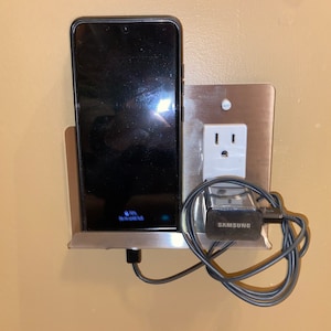 May include: A black smartphone is plugged into a Samsung charger, which is plugged into a wall outlet. The phone is mounted on a metal stand.