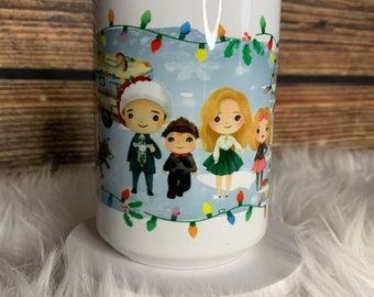 May include: White ceramic mug with a Christmas scene featuring a family of four standing in the snow. The family is dressed in winter clothes and there are Christmas lights strung up in the background.