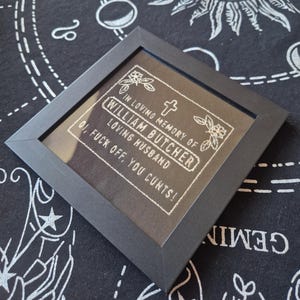 May include: A square black frame containing a black fabric panel with white embroidered text that reads: IN LOVING MEMORY OF WILLIAM BUTCHER LOVING HUSBAND OI, FUCK OFF, YOU CUNTS! The embroidery includes a small cross and floral accents.