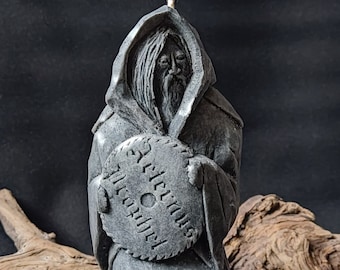 Op de afbeelding: Een grijze kaars in de vorm van een gekapte figuur die een ronde schijf vasthoudt met daarop gegraveerde runen. De kaars heeft een witte lont.