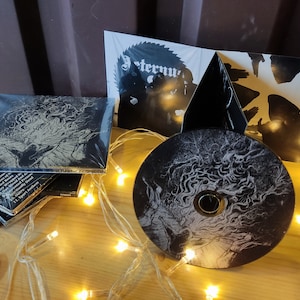 Op de afbeelding: Een zwarte CD met een wit en grijs ontwerp van draaiende lijnen. De CD is omringd door een string witte lampjes. De CD maakt deel uit van een muziekalbum getiteld "Eternum".