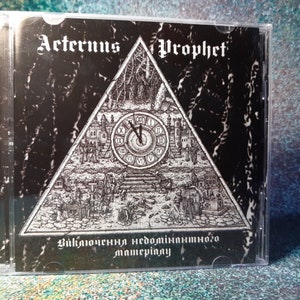 May include: A black CD case with a black and white image of a clock inside a triangle. The text "Aeternus Prophet" is at the top of the case. The text "Включение недоминирующего материала" is at the bottom of the case.
