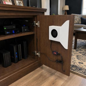 May include: A wooden cabinet with an open door revealing electronic components. Inside, there are several black devices and a white fan-shaped component. A small digital display shows the number 226. The cabinet is in a living room setting.