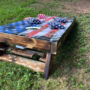 May include: A wooden cornhole game with an American flag design. The game is set up on a grassy surface with bean bags on top.
