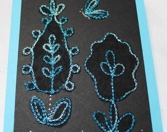 May include: A black card with a blue and white embroidered design of stylized flowers and a butterfly. The card is surrounded by a light blue border.