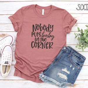 Peut inclure: Un t-shirt rose poudré avec l'inscription noire "Nobody puts baby in the corner". Il est associé à un short en jean et à des baskets Converse blanches. L'image est posée sur un fond en bois blanc.