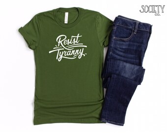 May include: A green t-shirt with the text "Resist Tyranny" in white lettering. The shirt is folded and displayed with a pair of blue jeans.