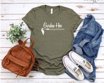 Può includere: Una maglietta verde con il testo "Garden Hoe" e un'illustrazione bianca di una zappa da giardino. La maglietta è su una superficie di legno bianco con uno zaino di pelle marrone, un paio di scarpe da ginnastica bianche e una giacca di jeans blu.