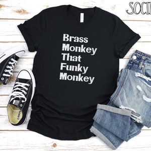 May include: Black t-shirt with white text that reads "Brass Monkey That Funky Monkey". The shirt is laid out on a white wooden surface with a pair of black trainers and blue jeans.