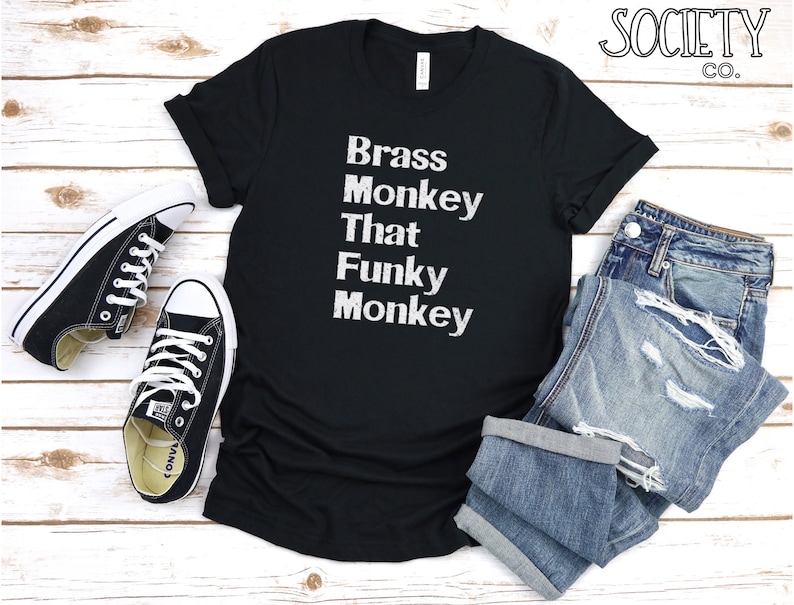 May include: Black t-shirt with white text that reads "Brass Monkey That Funky Monkey". The shirt is laid out on a white wooden surface with a pair of black sneakers and blue jeans.