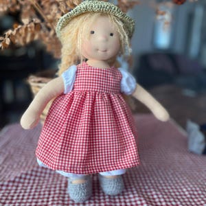 May include: A handmade doll with blonde hair wearing a red and white gingham dress and a straw hat. The doll is sitting on a red and white checkered tablecloth.
