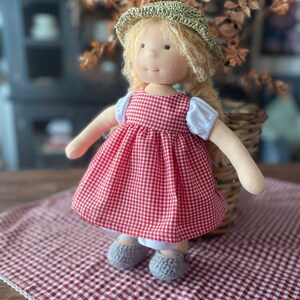 May include: A handmade doll wearing a red and white gingham dress, a straw hat, and gray crocheted shoes. The doll is sitting on a red and white gingham tablecloth.