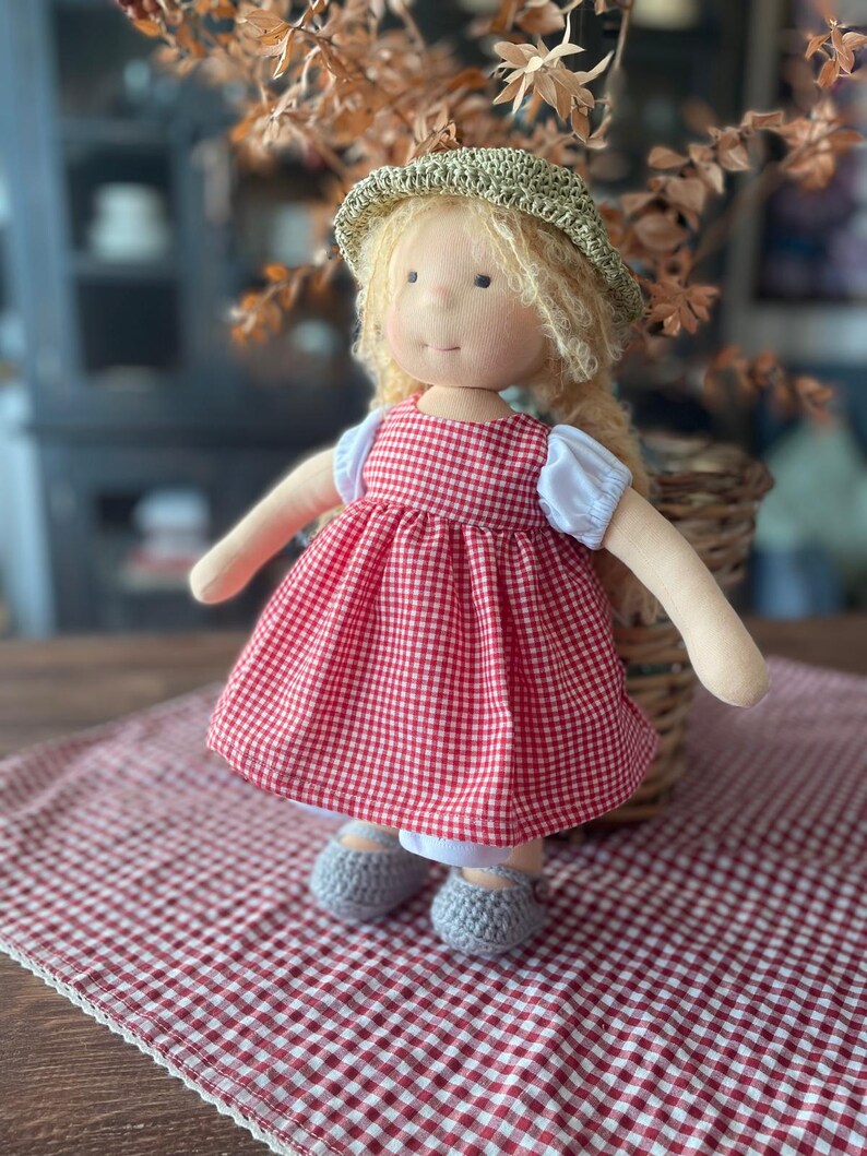 May include: A handmade doll wearing a red and white gingham dress, a straw hat, and gray crocheted shoes. The doll is sitting on a red and white gingham tablecloth.