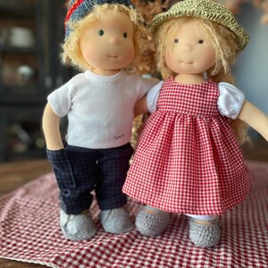 May include: Two handmade cloth dolls, one wearing a red and white gingham dress and a green hat, the other wearing a white shirt, blue pants, and a blue and red knit hat. The dolls are standing on a red and white checkered blanket.