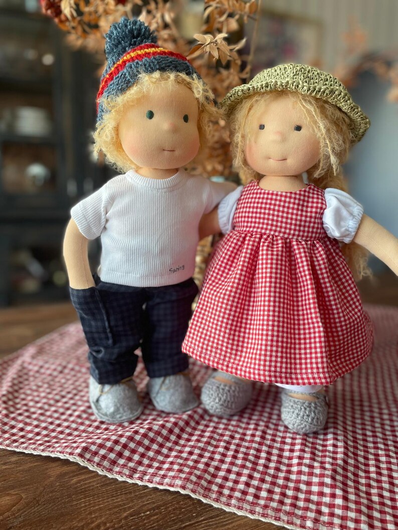 May include: Two handmade cloth dolls, one wearing a red and white gingham dress and a green hat, the other wearing a white shirt, blue pants, and a blue and red knit hat. The dolls are standing on a red and white checkered blanket.