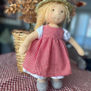May include: A handmade doll with blonde hair wearing a red and white gingham dress, a straw hat, and grey crocheted shoes.