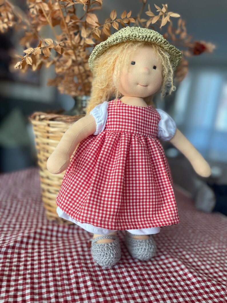 May include: A handmade doll with blonde hair wearing a red and white gingham dress, a straw hat, and grey crocheted shoes.