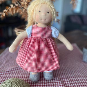 May include: A handmade doll with blonde hair wearing a red and white gingham dress. The doll is sitting on a red and white checkered tablecloth.