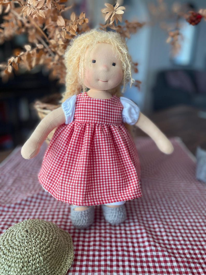 May include: A handmade doll with blonde hair wearing a red and white gingham dress. The doll is sitting on a red and white checkered tablecloth.
