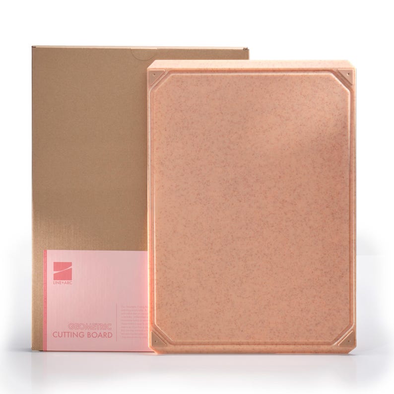 May include: A rectangular, speckled, peach-colored cutting board with a geometric design. The board is next to a cardboard box and a pink card with the text "GEOMETRIC CUTTING BOARD". The cutting board has a textured surface and decorative corner details.