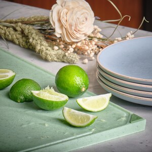 May include: A still life featuring fresh limes, some halved, on a light green cutting board. A stack of blue plates and a decorative flower arrangement are in the background. The limes are bright green and juicy.