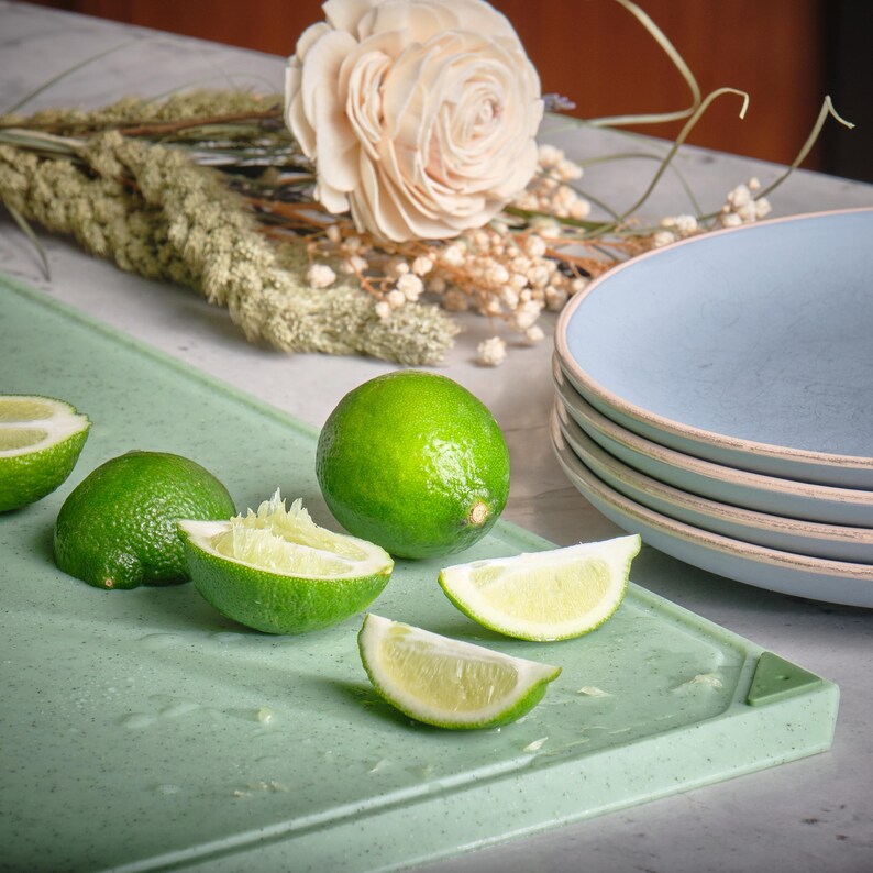 May include: A still life featuring fresh limes, some halved, on a light green cutting board. A stack of blue plates and a decorative flower arrangement are in the background. The limes are bright green and juicy.